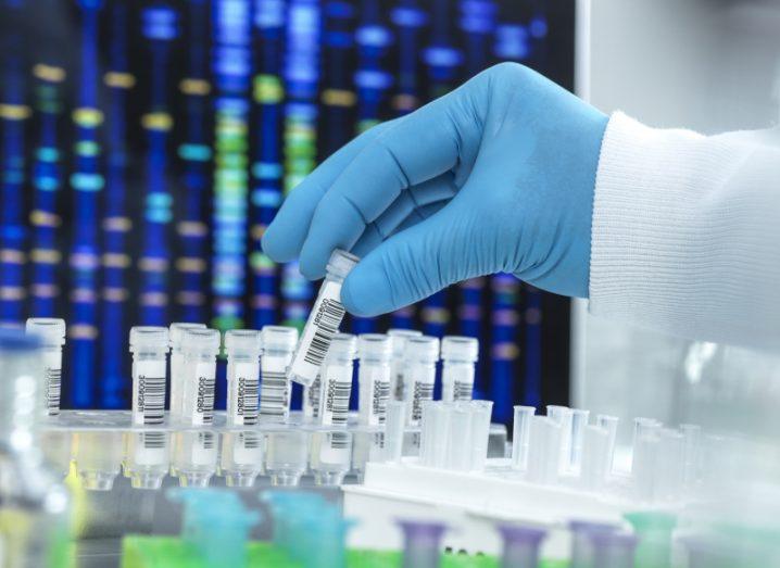 A hand in a blue protective glove selects a DNA sample from a row of test tubes. A genome sequence is visible on a screen in the background.