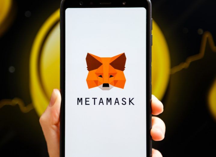 MetaMask logo on the white screen of a mobile phone with a cryptocurrency coin behind it.