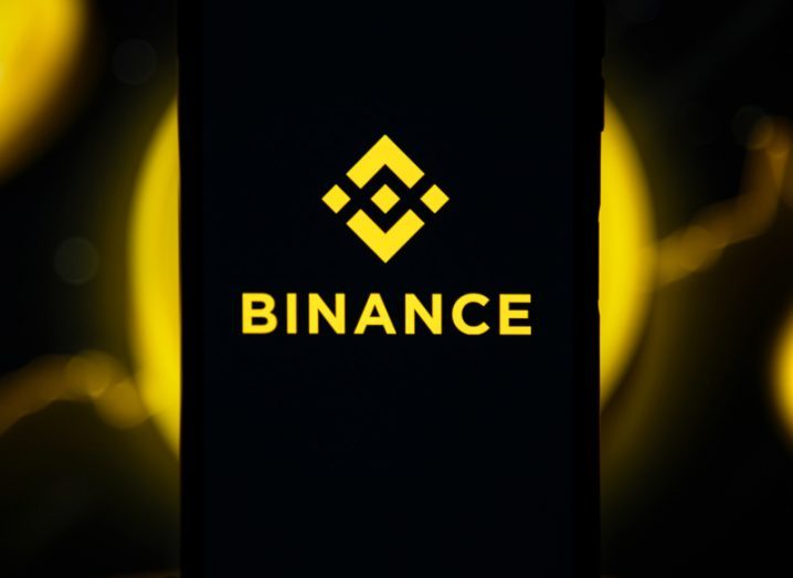 Binance name and logo on a dark mobile phone background with yellow lighting behind it.