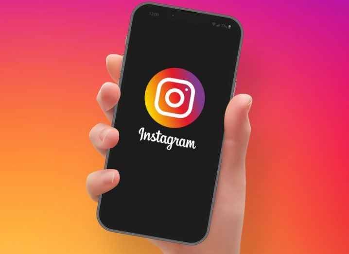 Hand holding a smartphone with the Instagram logo on its black screen against a pink and orange background.