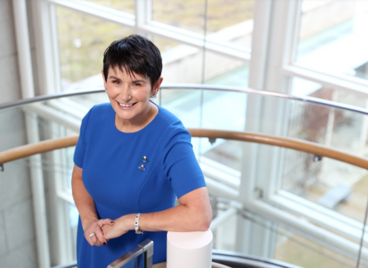 Carolan Lennon in a blue dress standing on a staircase beside a wall of windows looking out onto an office campus.