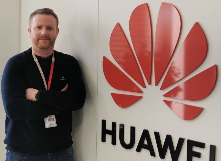 A man stands in front of the Huawei logo on a wall. He is smiling at the camera.