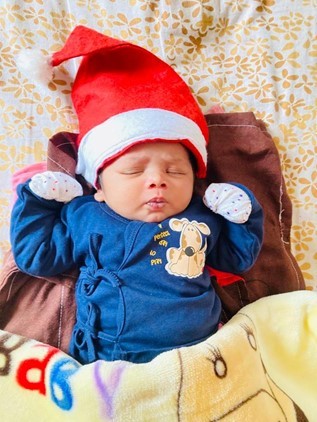 A tiny baby in a blue outfit and santa hat lies on the floor with his eyes closed.