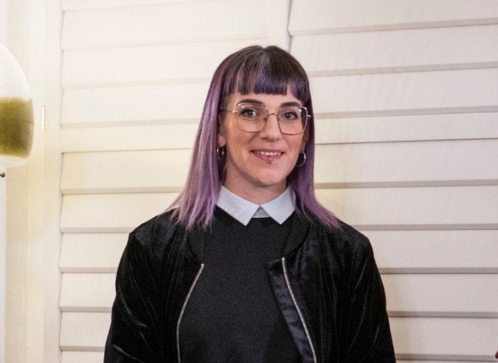 A woman with lilac hair and glasses smiles at the camera. She stands against a white wall.