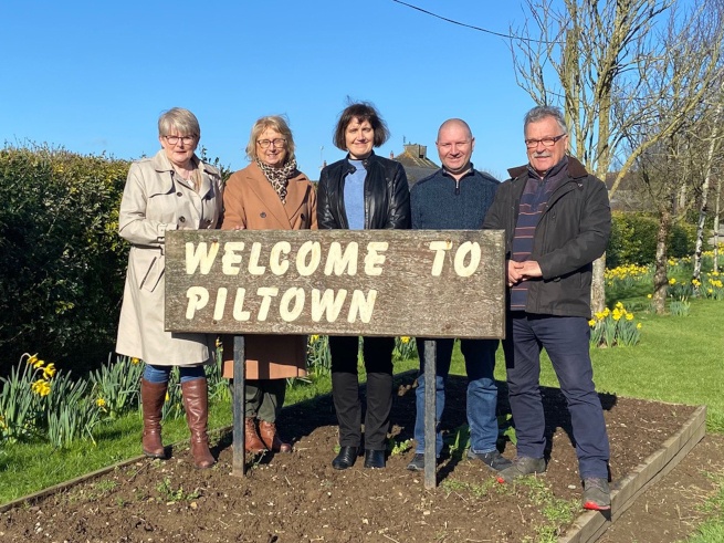 Three women and two men stand next to each other holding a sign that reads "Welcome to Piltown".