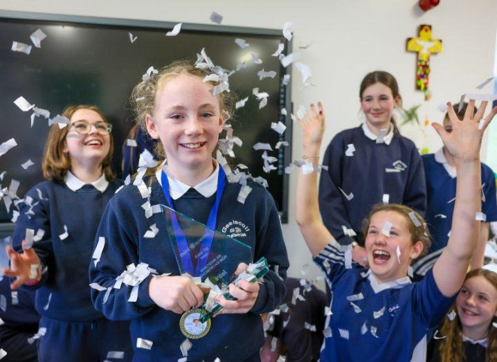 A girl wears the Intel Mini Scientist award medal and smiles at the camera while other girls around her celebrate amid a confetti shower.