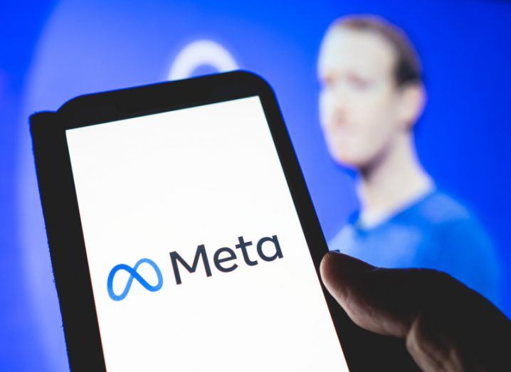 Meta logo on a smartphone held in a hand with blurred image of Meta CEO Mark Zuckerberg in the background.