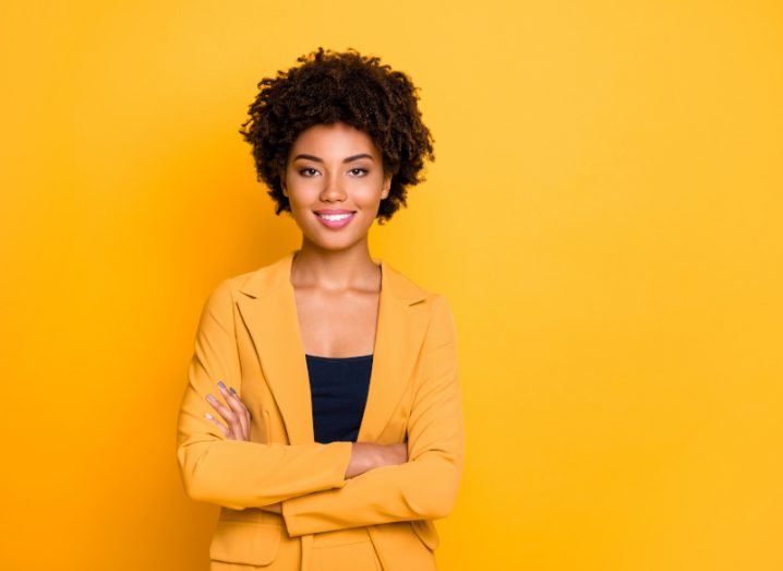 A mixed-race woman stands with folded arms in a bright yellow background.