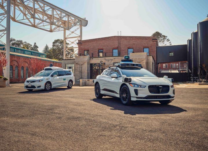 Two white Waymo SUVs with sensors on top of them parked next to each other on a dirt road.