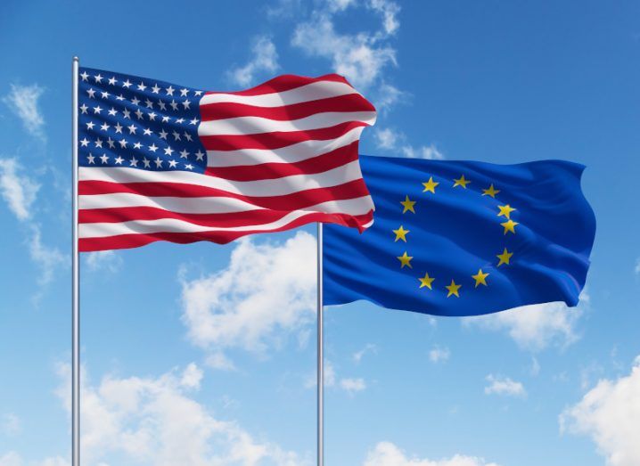 The US and EU flags on poles next to each other with a blue sky above.