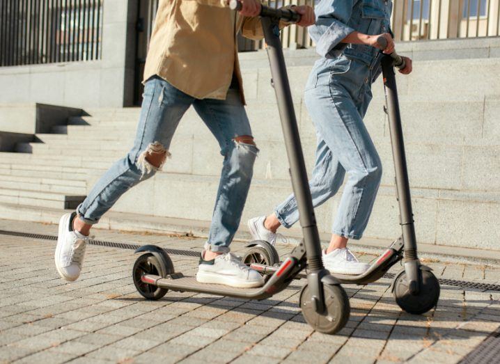 Two people on electric scooters in a city on a sunny day.