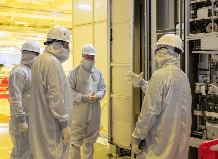 Four people wearing white factory outfits and helmets talking to each other inside a manufacturing plant.