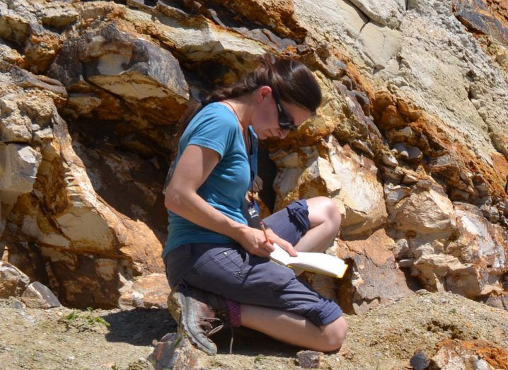 A woman sitting on the ground taking notes surrounded by a rocky area. She is researching fossil records.
