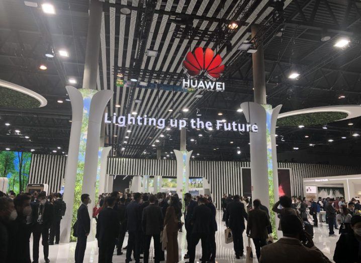 Huawei logo with the phrase "lighting up the future" below it, with people attending the Huawei booth at MWC 2022.