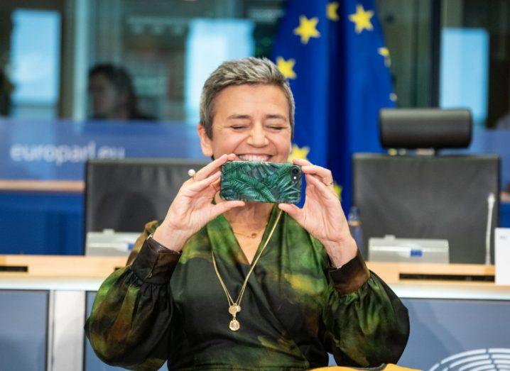 Wearing a moss-green long-sleeved dress, Margrethe Vestager sits at the benches of European Parliament in front of an EU flag, smiling as she holds up her smartphone to take a photograph.