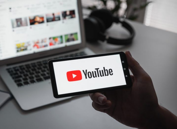 Youtube logo on a mobile phone screen, being held by a person's hand with a laptop and a table in the background.