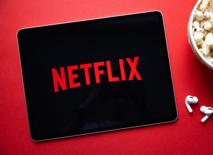 Netflix logo on a mobile tablet screen lying on a red table with wireless earphones and a bowl of popcorn next to it.
