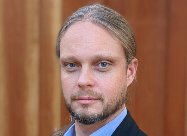 A headshot of Chris Evans from HackerOne. He is wearing a dark suit and standing against an out of focus brown wall.