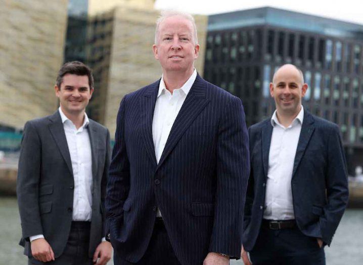 Three men stand in a row, with two slightly further back. They are all wearing suits and standing near Grand Canal Dock.