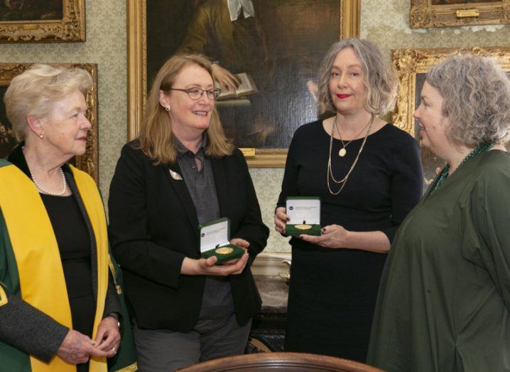 Four women standing in a group at an awards ceremony held by the Royal Irish Academy in Trinity College Dublin. Paintings in gold frames on a pale green wall in the background.
