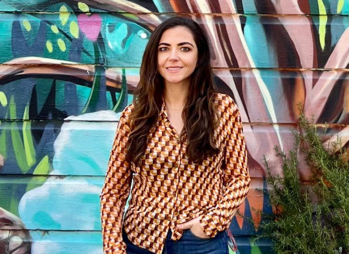 A young woman with long brown hair smiles at the camera. She is wearing a patterned shirt and standing in front of a graffiti wall.