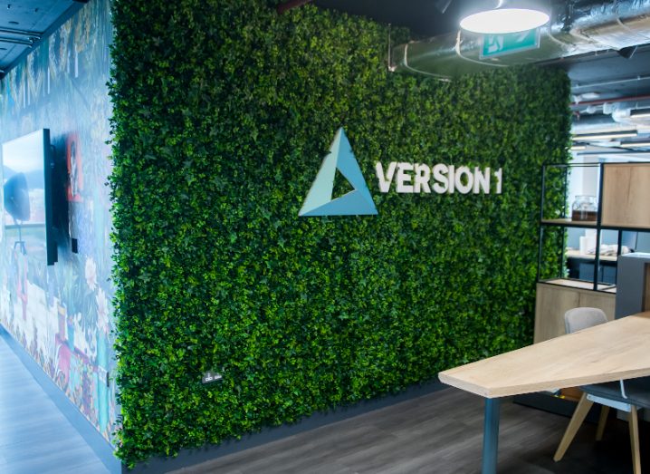 The Version 1 logo on a wall of artificial greenery in a bright office.