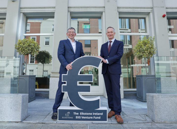 Two men in business formals stand on either side of a big euro sign.