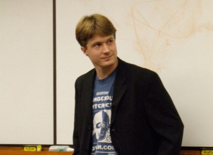 Photo of Virgil Griffith wearing a black jacket over a t-shirt and standing in front of a white board.
