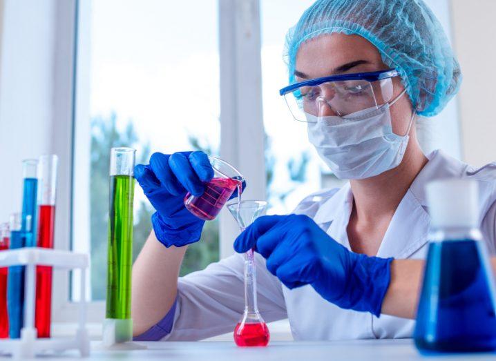 A lab worker wearing a mask pours a liquid from one vial to another.