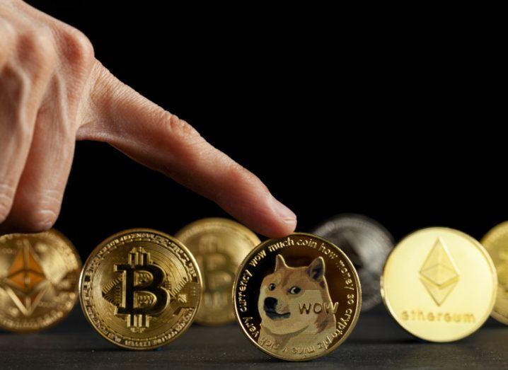 Several different crypto coins stand upright against a black background while a hand points to one in the centre with a dog on it.