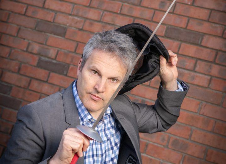 Headshot of Patrick Dight, the founder of Sparwatch, holding a fencing sword with a brick wall behind him.
