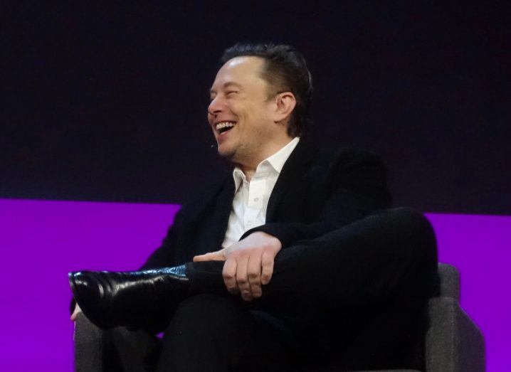 Photograph of Elon Musk laughing while sitting on a chair, with a hand on his leg.