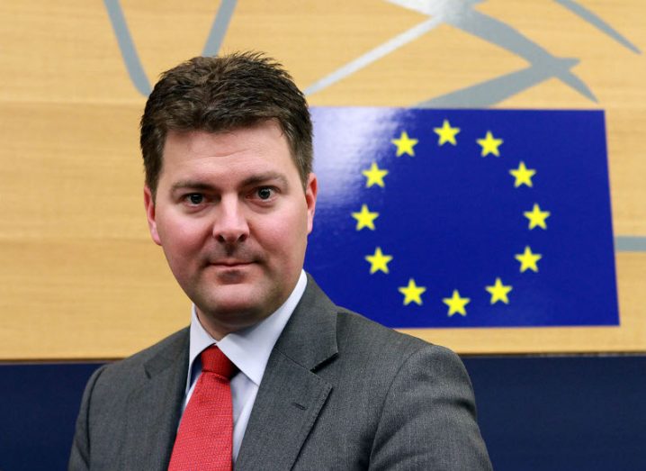 Photograph of a man in a grey suit with a red tie looking at the camera, with the EU flag behind his head. He is Andreas Schwab MEP.