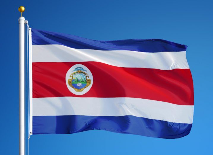 Photograph of the flag of Costa Rica, waving on a flag pole with a blue sky behind it.
