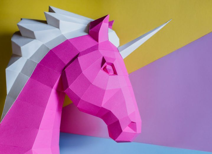 A geometric paper unicorn bust in pink and white against a background of yellow, purple and blue.
