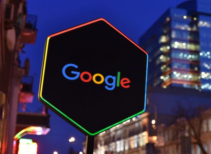 Google logo on a hexagonal sign with a black background and different coloured lighting around it, with city buildings behind the sign.