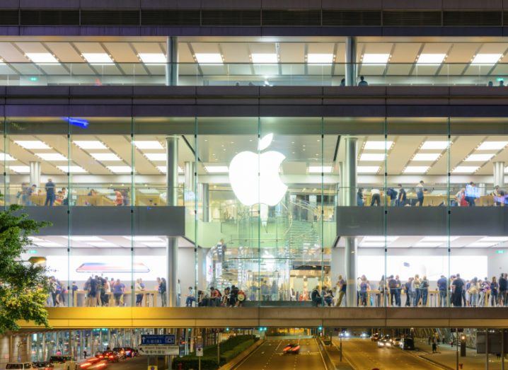 Apple logo in front of a store with people inside at night. The building is above a road where cars are driving.