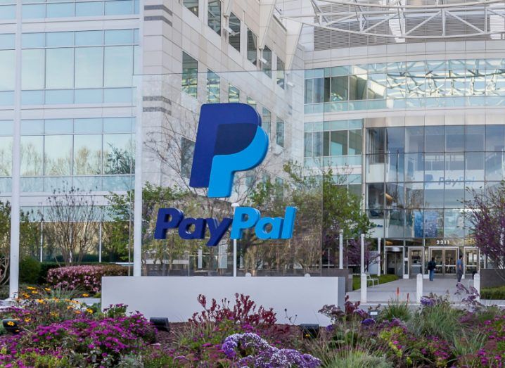 Photograph of the PayPal company logo on a glass pane in front of a large building. There are flowers and grass around the ground of the glass pane.