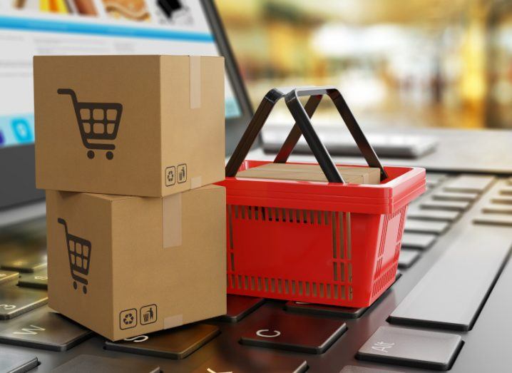 Cardboard shipping boxes with shopping trolley symbols on them, next to a red shopping basket that is on a laptop keyboard, with the laptop screen visible.