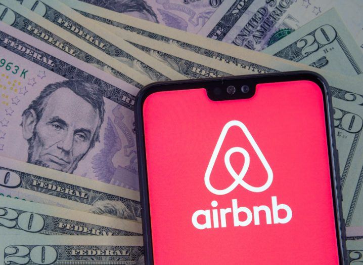 A smartphone with the Airbnb logo on the screen, lying against a background of US dollar bills.