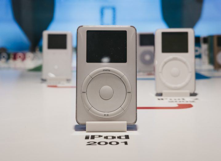 A range of iPods laying on a table with red lines connecting them, to show a timeline of the product. The original iPod model from 2001 is at the centre of the image, with later versions of the iPod behind it.