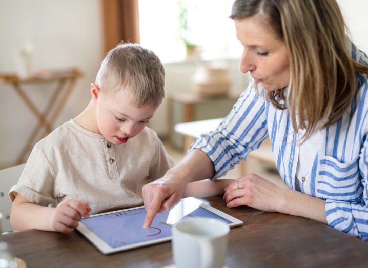 A little boy with Down's Syndrome of primary school age sitting at a table using a tablet with assistive technology supervised by a woman.
