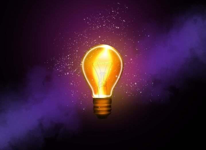 Lit-up light bulb with some sparks of light around it, in a dark background with a purple cloudy shade diagonally in the background.