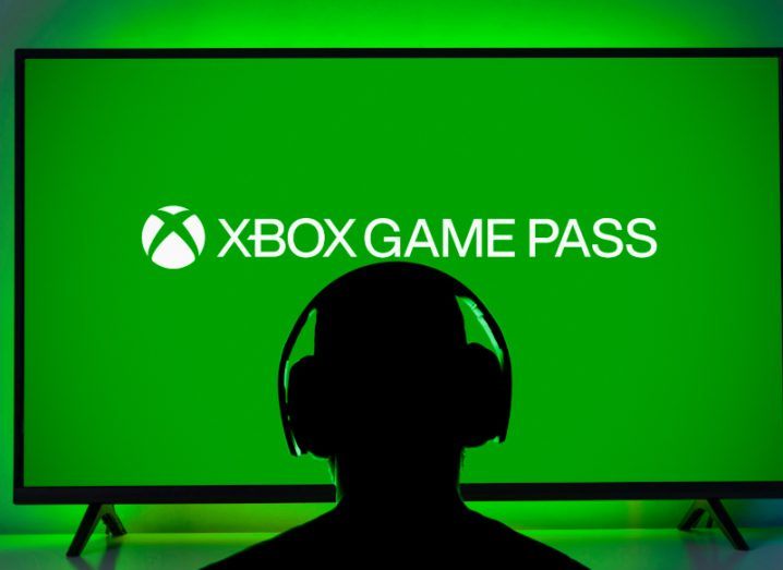 Photograph of a person wearing headphones, looking at a TV that has Xbox Game Pass written on it against a green background.