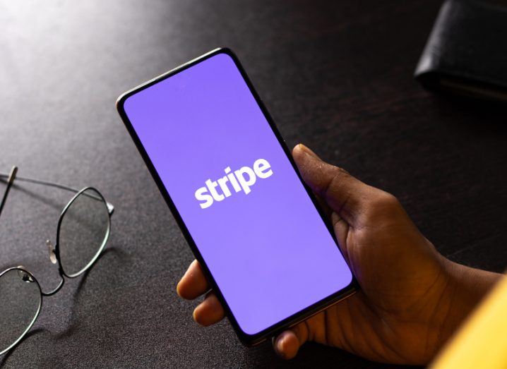 Stripe logo in a purple background on a mobile phone, being held in a person's hand. There is a dark table under the phone with a pair of glasses resting on it.
