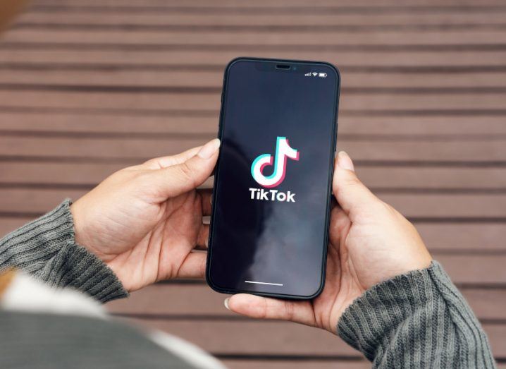Person holding a mobile phone in both hands that has the TikTok logo on it, with a wooden floor in the background.
