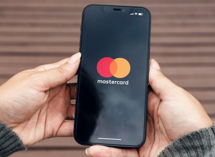 Mastercard logo on a mobile phone, being held in a person's hands with a wooden ground as the background.