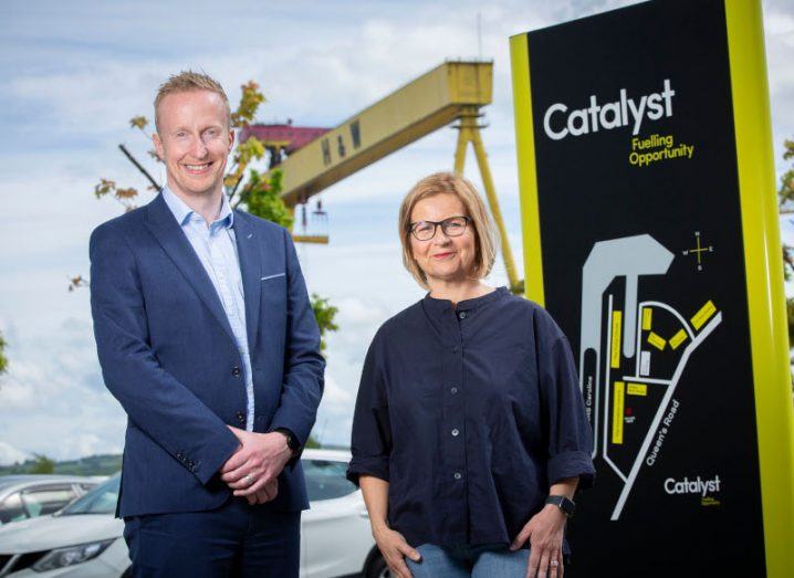Photograph of a man and a woman smiling for the camera with a black and yellow sign next to them with the Catalyst name on it. They are Niall Devlin and Elaine Smyth.