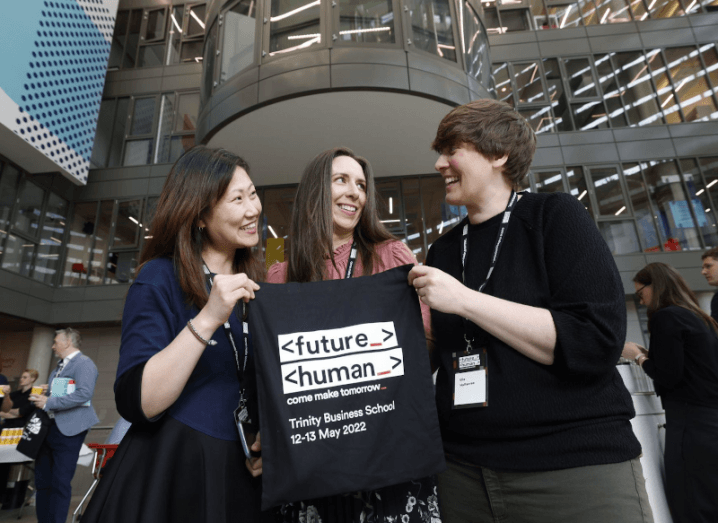 Three women stand outside the entrance to Trinity Business School, holding up a tote bag with the Future Human logo.