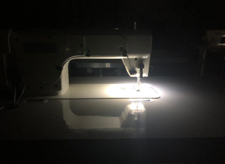 A sewing machine in a dark room, with the light of the machine showing the needle. The machine is on a grey table.